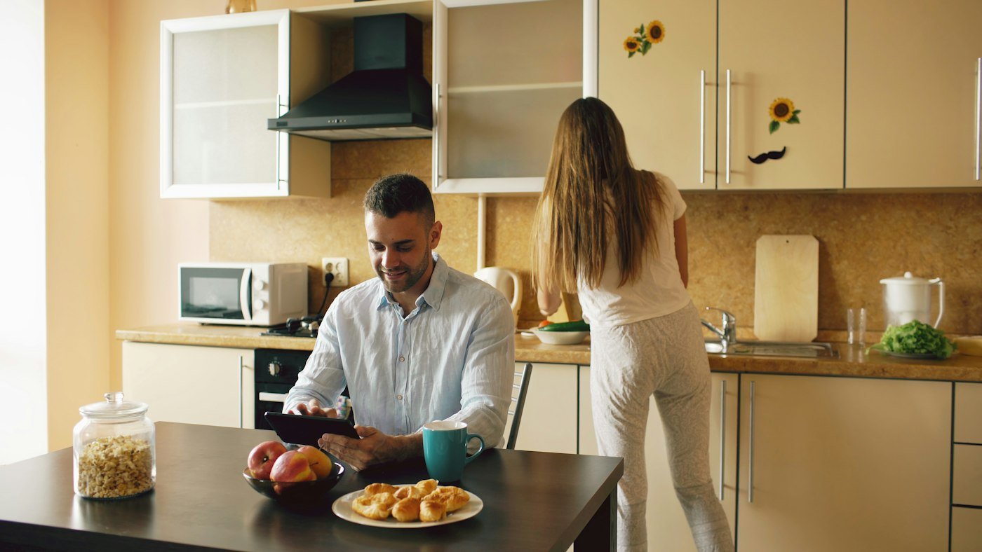 Couple in the kitchen, he is on a tablet while she cooks